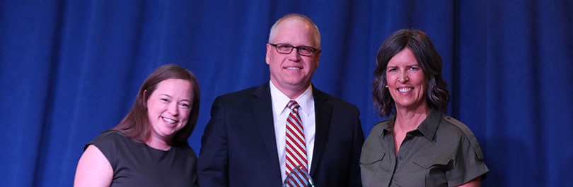 Two smiling women and 1 smiling man dressed up for an event stand in front of a blue curtain background.