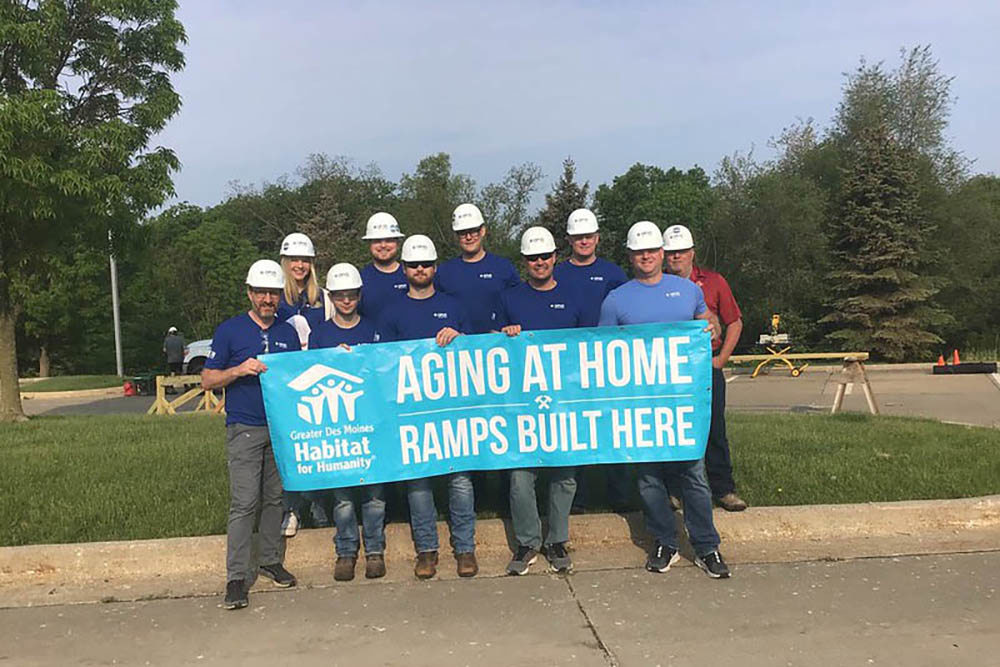 group of men and women wearing blue shirts and hard hats and holding a banner reading "Aging at Home Ramps Built Here"