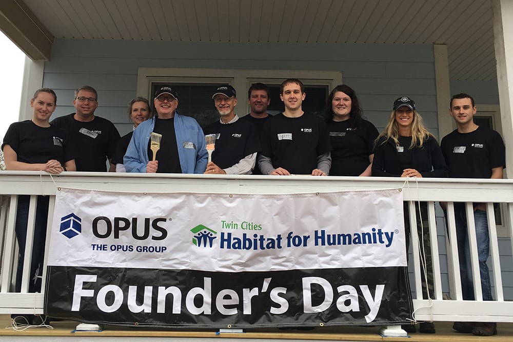 group of men and women on house porch with banner reading "The Opus Group, Twin Cities Habitat for Humanity, Founder's Day" on porch railing