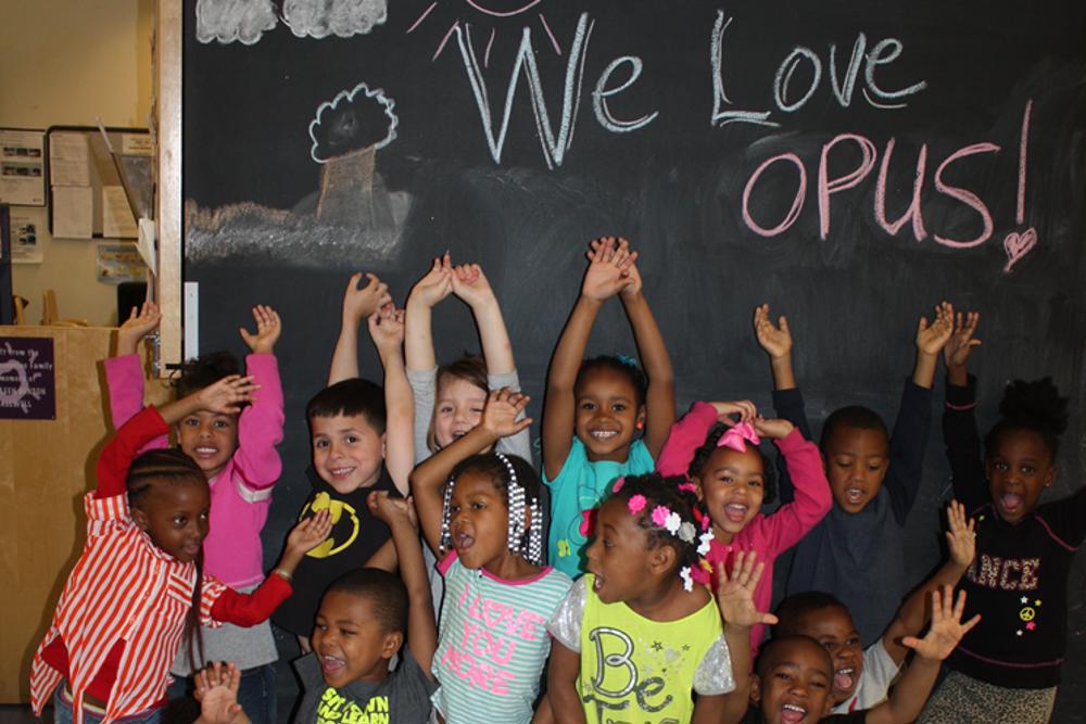 a group of children with arms held up in front of a chalkboard that says, "We Love Opus!"