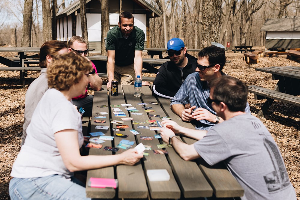 men and women playing a card game on a picnic table in a wooded picnic area