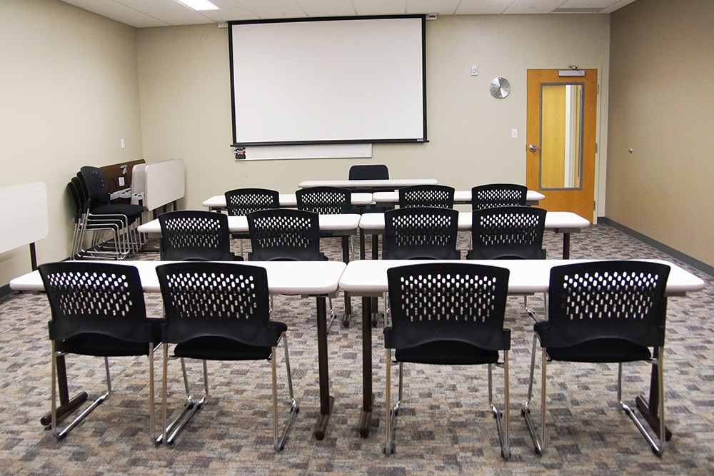 meeting room in an institutional building with rows of tables with chairs in the foreground facing a dry erase board in the background