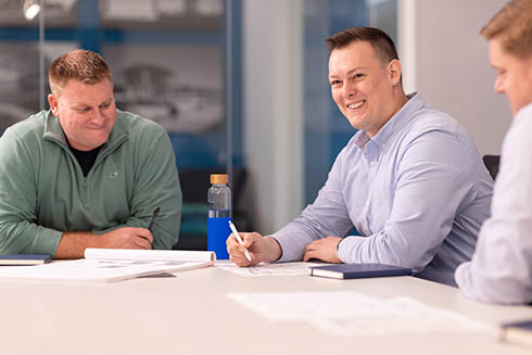 3 men in professional attire sit at a table smiling while collaborating on work.