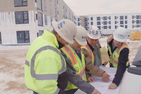 A group of people in high-visibility safety gear stand in front of a construction site and discuss building plans.