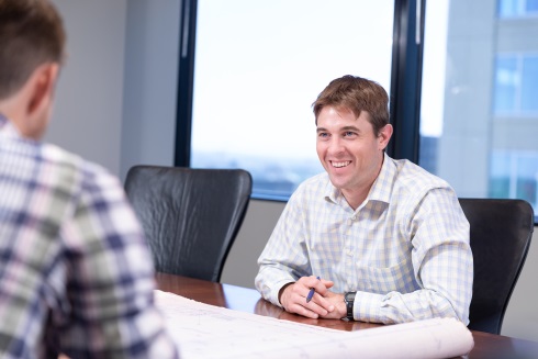 A smiling man in professional attire sits at a conference room table across from another person.