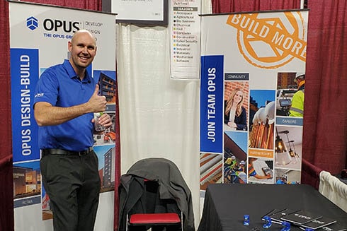 A smiling man in front of banners at a career fair gives a thumbs up.