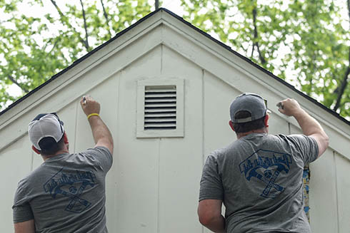 Two men paint the exterior of a home with Building Community on the back of their shirts.