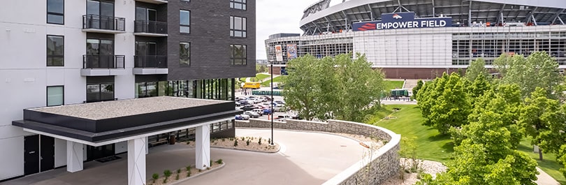 The front entrance to a new apartment building with a football stadium and healthy trees in the background.