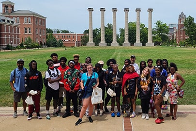 A large group of young people pose for a photo in front of a lawn on a college campus.