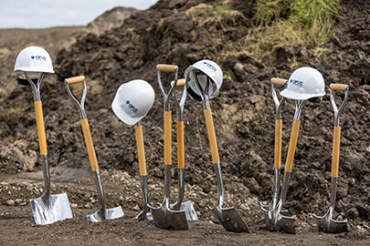 9 shovels stand upright in bare dirt with hard hats on some and a large pile of newly turned dirt in the background.