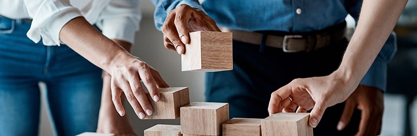 Three diverse people reach forward and stack wooden blocks on a table in front of them.