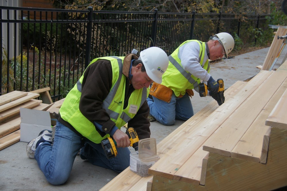 two men wearing safety vests and hard hats kneeling at the bottom of wooden stairs with drills fastening wood to the bottom stair