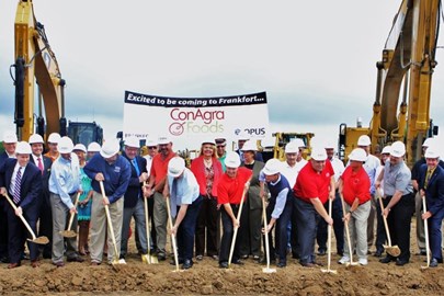 a group of men and women wearing hard hats with front row of people shoveling dirt in foreground and sign reading "Excited to be coming to Franfort...ConAgra Foods" and heavy construction equipment in background