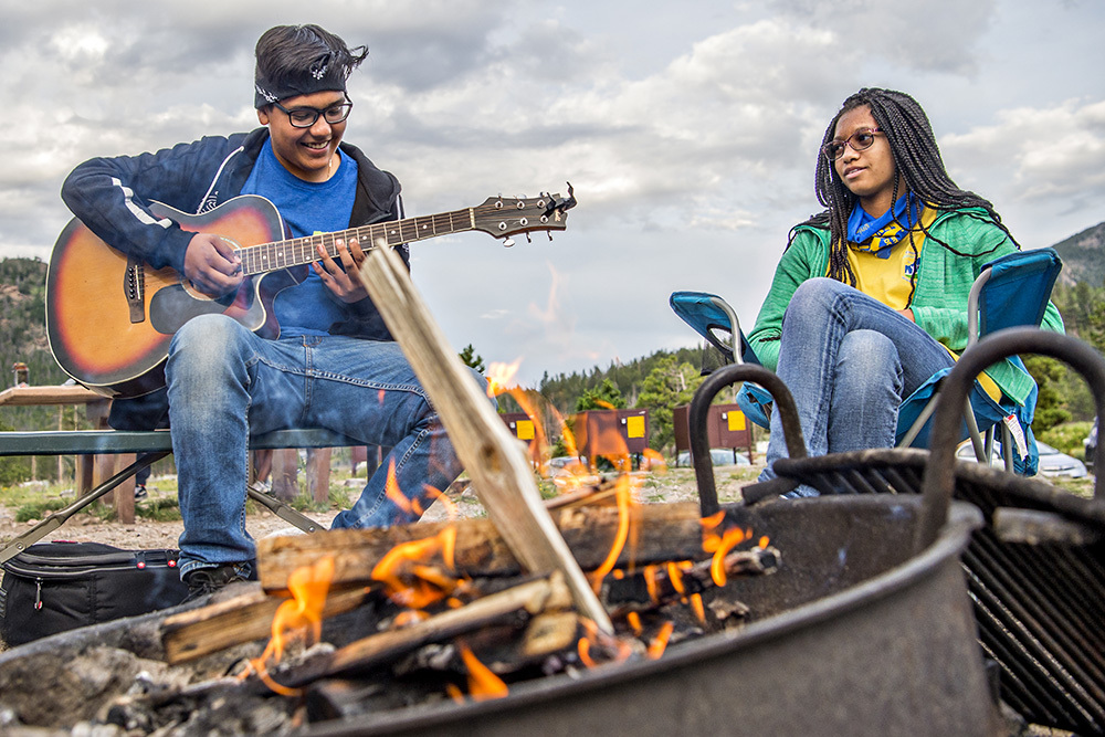 Boy with guitar and girl sitting around campfire
