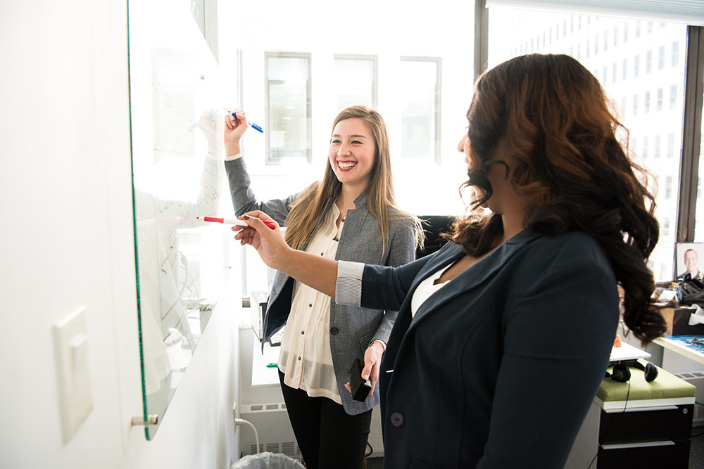 two women in professional attire writing on a whiteboard in a conference room
