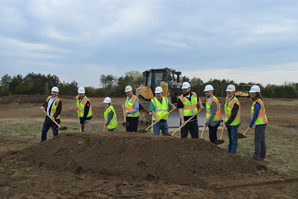 Group of men and women wearing safety vests and hardhats with shovels throwing dirt at a construction site groundbreaking