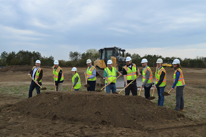 Group of men and women wearing safety vests and hardhats with shovels throwing dirt at a construction site groundbreaking