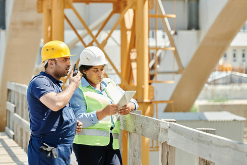 Man and woman in hardhats at construction site with man talking into walkie-talkie and woman looking at a paper in her hands