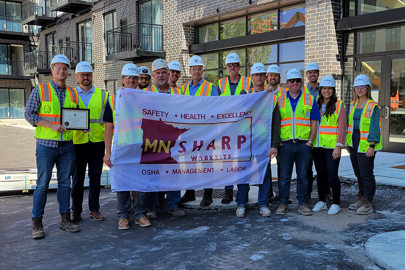 Group of men and women wearing safety vests and hardhats with MNSHARP banner and framed certificate in front of a residential apartment building
