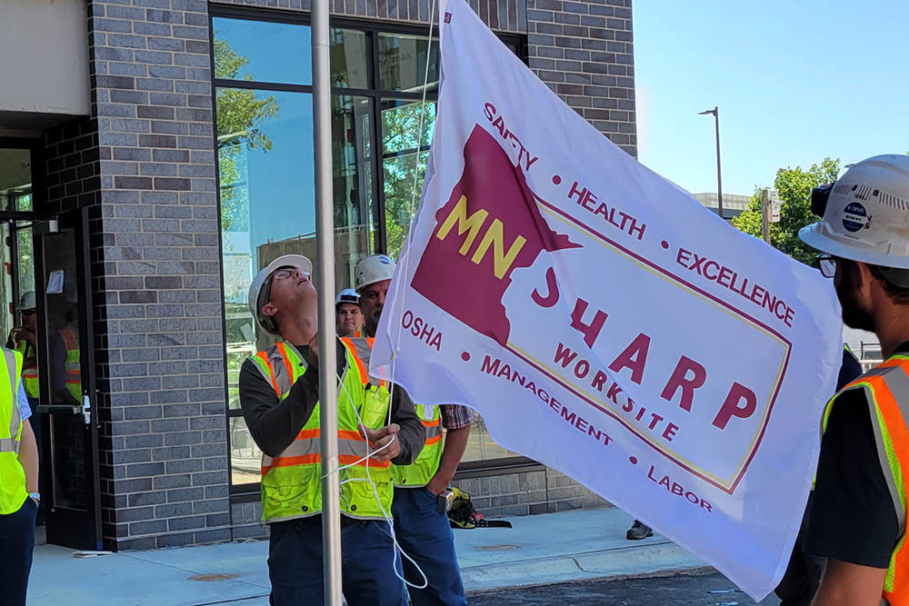 man in hardhat and safety vest raising a MNSHARP flag outside of an apartment building