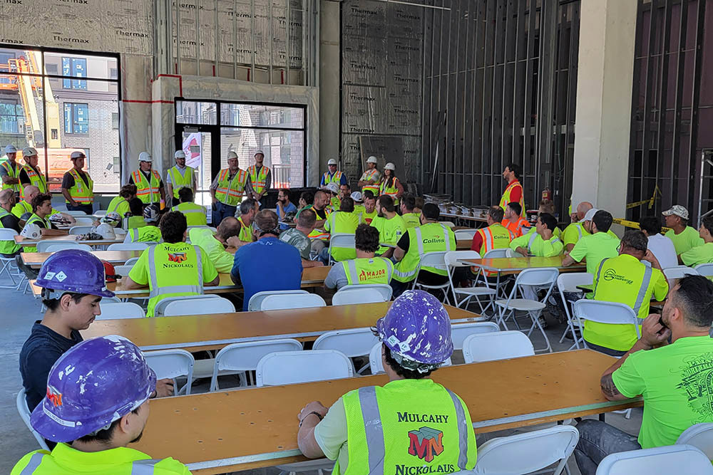 inside an unfinished building, men and women wearing hardhats and safety vests standing and presenting in the background and men and women in safety vests seated in foreground