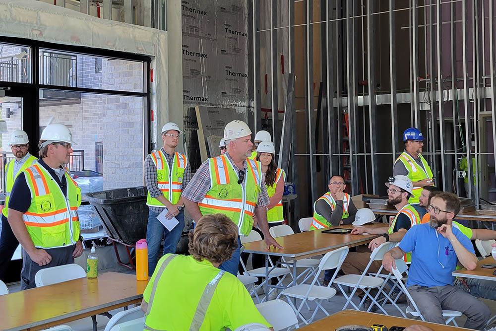 man wearing hardhat and safety vest talking to group of men and women wearing hardhats and safety vests inside unfinished apartment building