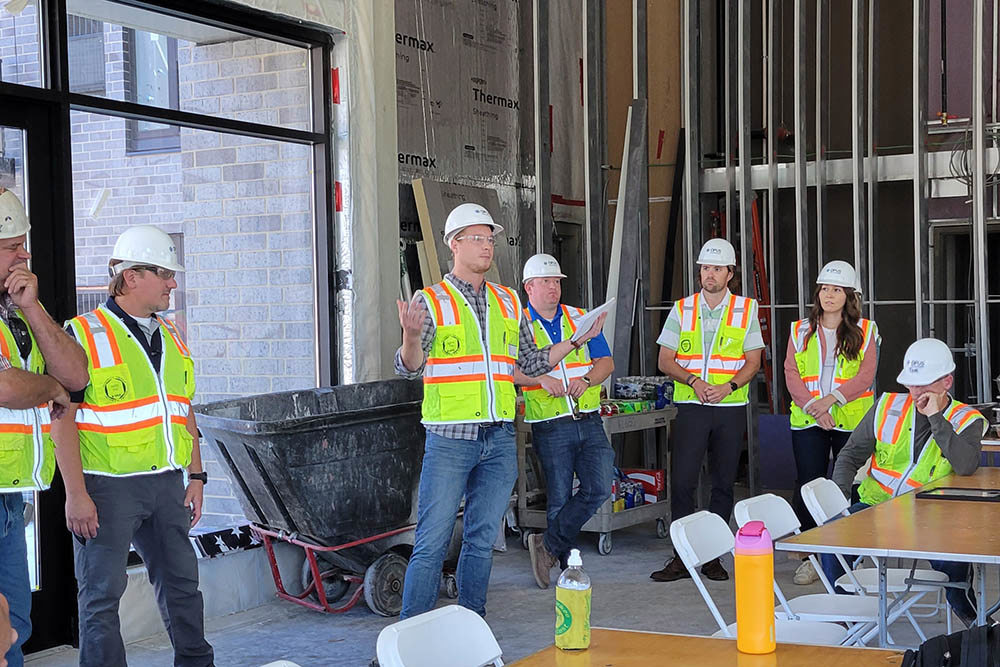 man wearing hardhat and safety vest talking to group of men and women wearing hardhats and safety vests inside unfinished apartment building