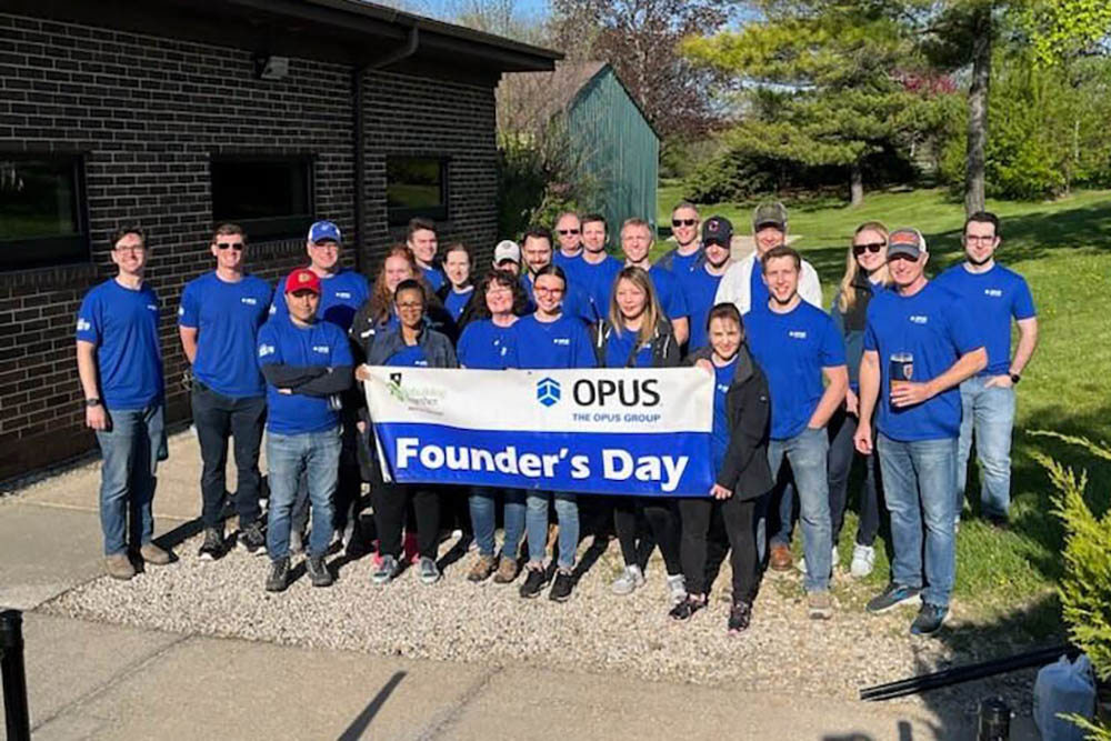 group of men and women wearing blue shirts and holding a banner reading "The Opus Group Founder's Day"