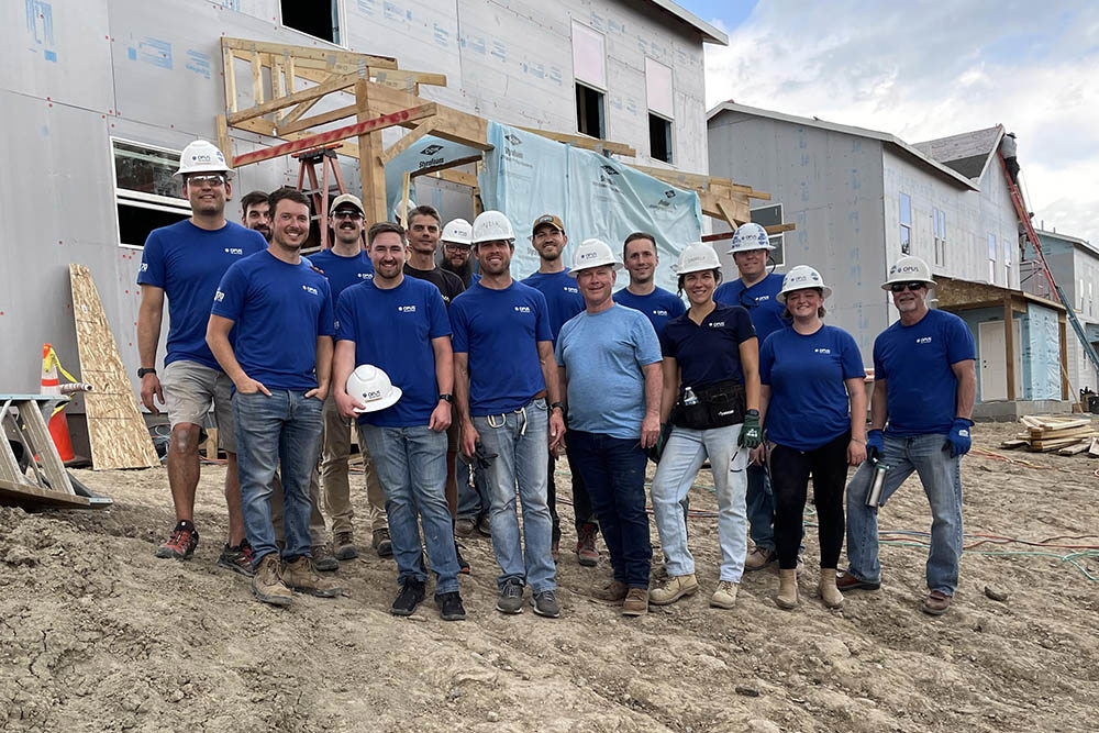 group of men and women wearing hard hats and blue shirts standing in front of a house under construction