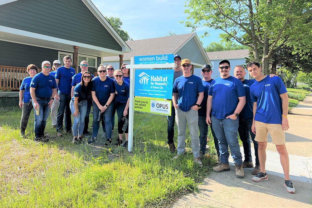 men and women wearing blue shirts standing outside next to a sign reading "Habitat for Humanity" and "The Opus Group" with a house in the background