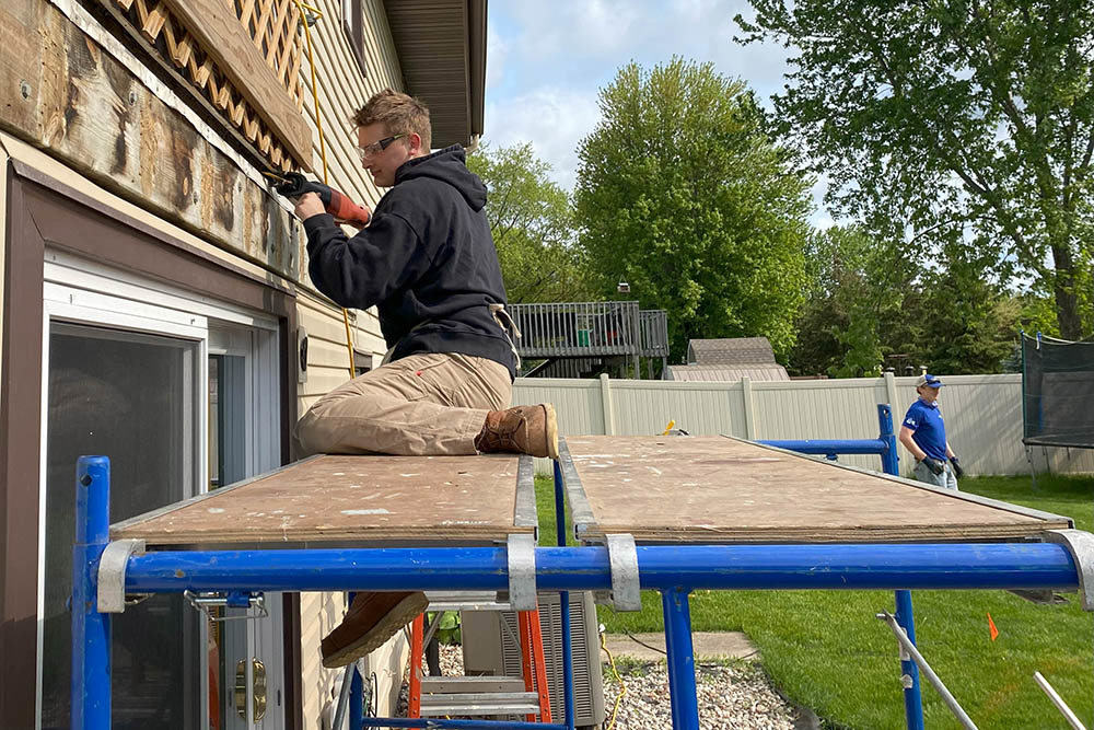 man kneeling on scaffolding while working on the siding of a house