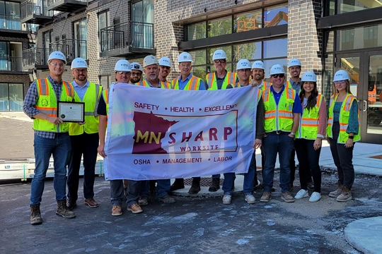 Group of men and women wearing safety vests and hardhats with MNSHARP banner and framed certificate in front of a residential apartment building
