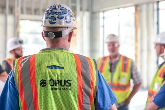 back of man wearing hardhat and safety vest with the words "The Opus Group" in the foreground and three men wearing hardhats and safety vests in background
