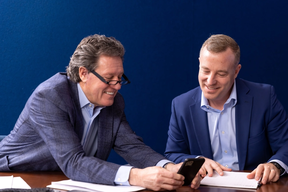 two men in professional attire looking at a phone and talking while seated at a table
