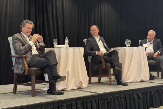 three men wearing professional attire sitting on a stage speaking
