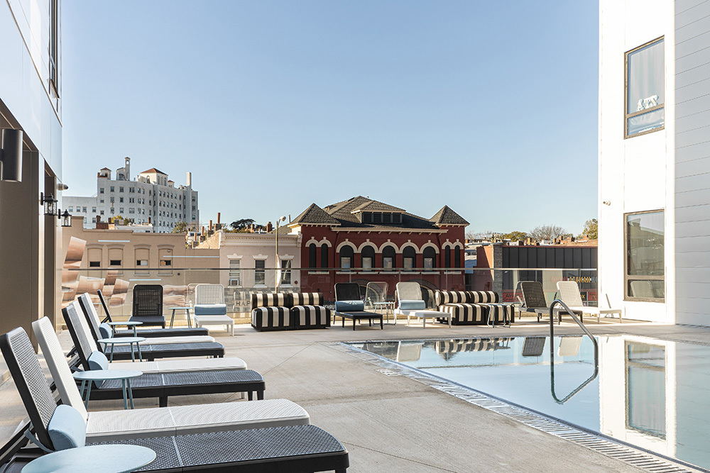 a rooftop outdoor living area in an apartment building with a row of lounge chairs on the left and a swimming pool on the right and chairs in the background