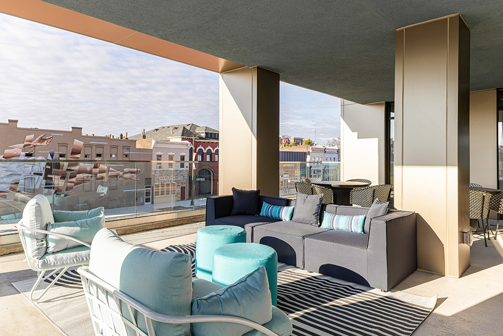 a rooftop outdoor living area in an apartment building with a covered seating area with ottomans surrounded by chairs and a sectional couch in the foreground and a table and chairs in the background