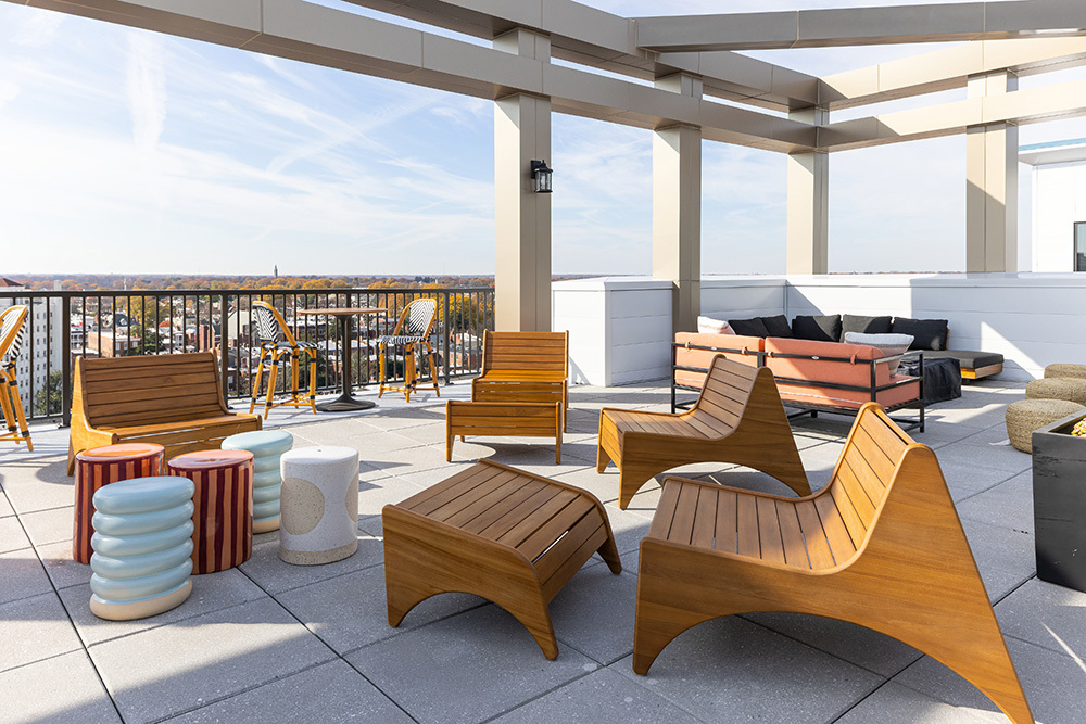 a rooftop outdoor living area in an apartment building with wooden chairs and footstool and stools in the foreground and a seating area and tables and chairs along the railing in the background