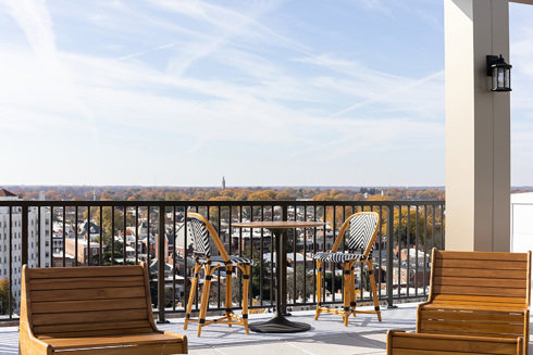 a rooftop outdoor living area in an apartment building with wooden chairs in the foreground and a table with two chairs along the railing in the background