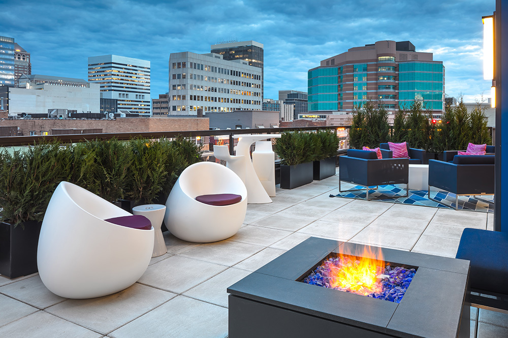 rooftop outdoor living area of an apartment building with a firepit and chairs in the foreground and a city view in the background