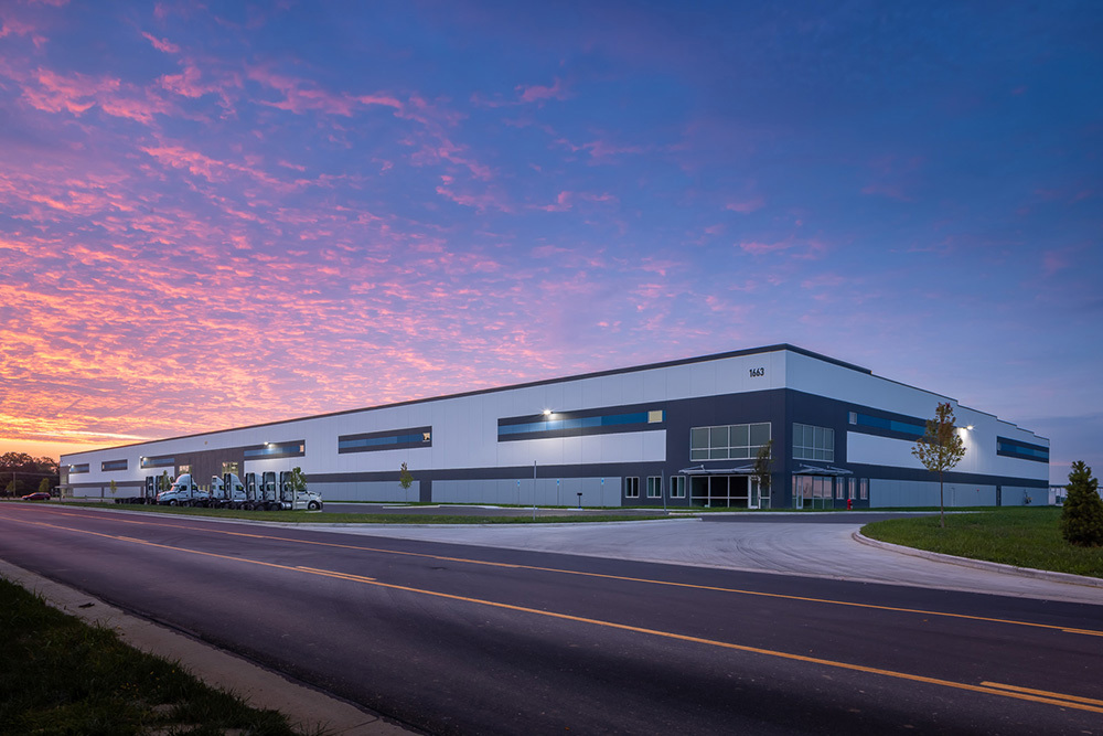 Multiple front entrances of a multi-tenant speculative industrial building taken from across the road at dusk with multiple semis