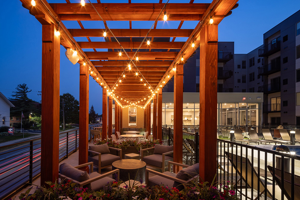 sunset view of rooftop outdoor living area of an apartment building with seating areas under lit pergola with lounge chairs and street on left and swimming pool on right