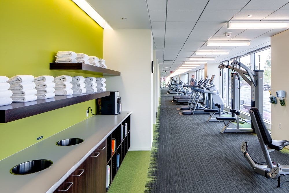 fitness center in an office building with base cabinets and shelves of towels on the left and exercise equipment along a wall with windows on the right
