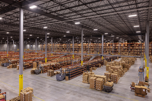 warehouse in an industrial building with rows of short shelving filled with boxes and conveyor belt in foreground and filled tall shelving in background