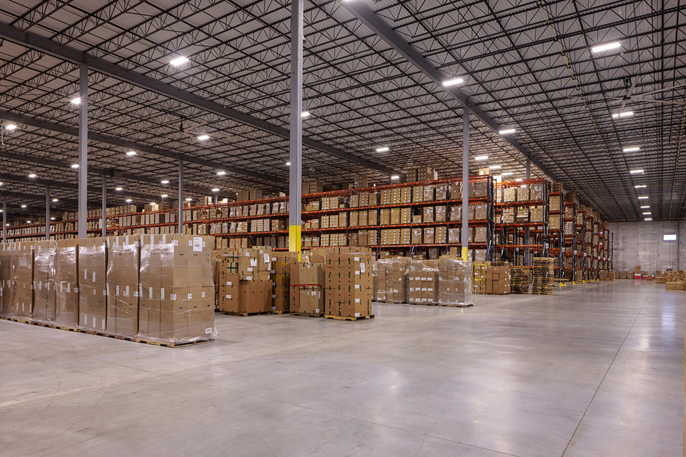 warehouse in an industrial building with rows of short shelving filled with boxes in foreground and filled tall shelving in background
