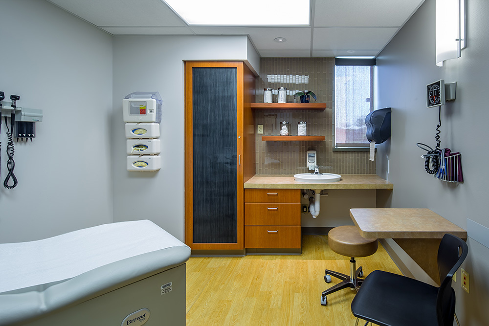 medical exam room in a healthcare building with exam table on left and small table with chair and stool on right in foreground and closet, sink, cabinet and shelves in the background