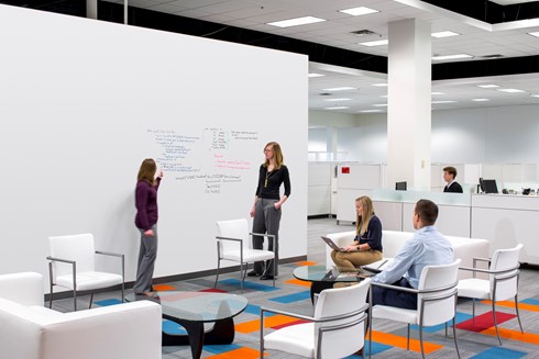 collaboration space in an office building where a man and woman sit facing two women who are writing on a dry erase board in the foreground and cubicles are in the background