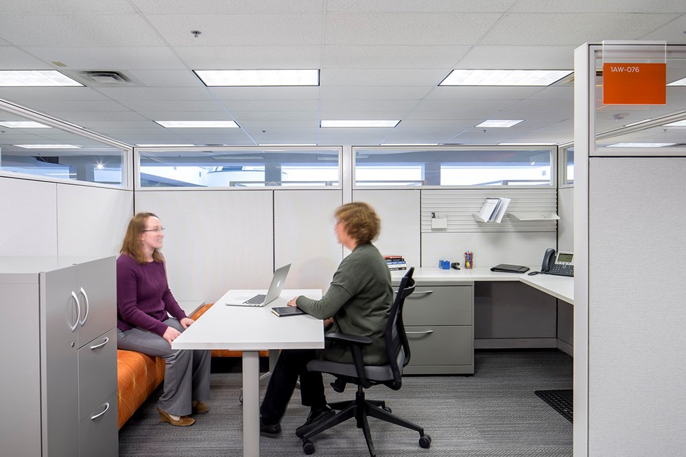 cubicle office in an office building where two women sit across from each other at a table in the center of the cubicle with a filing cabinet in the foreground and corner desk in the background