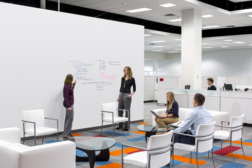 collaboration space in an office building where a man and woman sit facing two women who are writing on a dry erase board in the foreground and cubicles are in the background