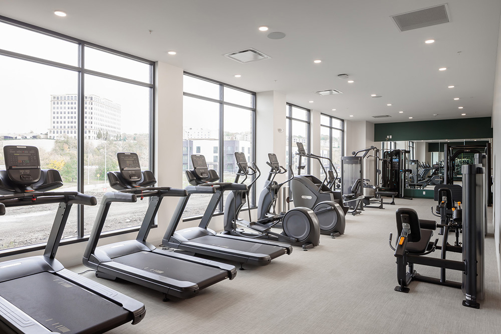fitness center in an apartment building with a row of exercise equipment along an exterior wall with windows